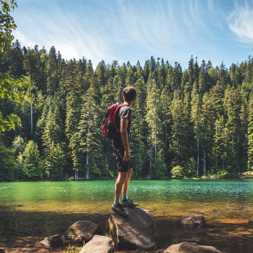 Hiker facing a lake surrounded by fir trees in the Vosges, starting from Les Bas Rupts hotel